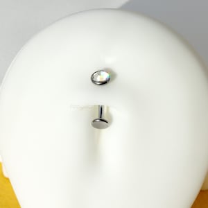 Puede incluir: Una barra de plata con una gema transparente iridiscente en un extremo y una bola plana de plata en el otro extremo. La barra está en una cabeza de maniquí blanca.