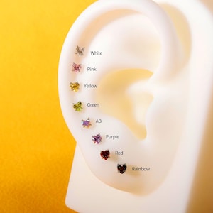 May include: A selection of stud earrings in various colors and shapes, including butterflies and hearts. Colors include white, pink, yellow, green, AB, purple, red, and rainbow. The earrings are displayed on a white ear model with color labels.