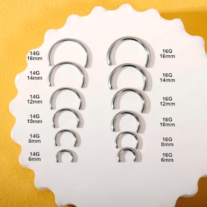 May include: A collection of silver-colored metal circular barbell rings arranged in two vertical columns. The left column lists sizes 14G from 6mm to 16mm, and the right column lists sizes 16G from 6mm to 16mm, with each size clearly labeled in black text.