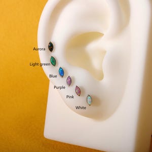 May include: A display of seven marquise-shaped earrings in various colors: aurora, light green, blue, purple, pink, and white. The earrings are set on a white ear model, showcasing their design and color variations.
