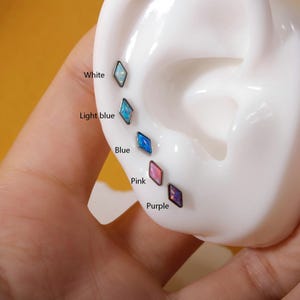 May include: A close-up of a white ear model with multiple diamond-shaped earrings in various colors: white, light blue, blue, pink, and purple. The earrings are labeled with their respective colors.