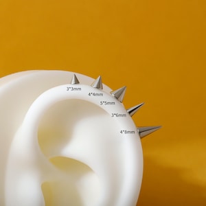 May include: A close-up of a white ear model with silver-colored spike earrings. The earrings are various sizes, including 3*3mm, 4*4mm, 5*5mm, 3*6mm, and 4*8mm. The background is a solid orange color.