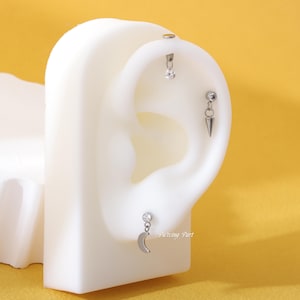 Può includere: Tre piercing per orecchie argentati su un modello di orecchio bianco. Il piercing superiore ha una piccola pietra trasparente, il piercing centrale ha una piccola pietra trasparente e un cono argentato, e il piercing inferiore ha una piccola pietra trasparente e una mezzaluna argentata.