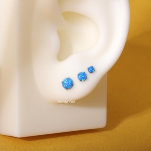 May include: Three blue opal earrings in different sizes, shown on a model's ear. The earrings are set in a silver tone metal.