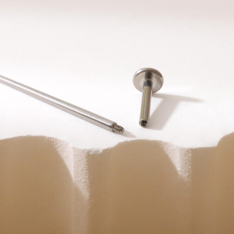 May include: A close-up of a silver barbell piercing with a flat, round top. The barbell is separated from its post, which is also silver.