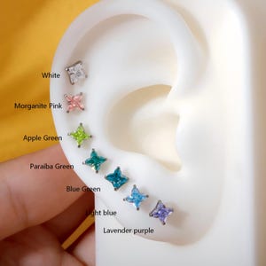 May include: A selection of star-shaped stud earrings in various colors, including white, pink, green, and blue. The earrings are displayed on a white ear model, with color names labeled: White, Morganite Pink, Apple Green, Paraiba Green, Blue Green, Light Blue, and Lavender Purple.