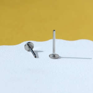 May include: Two silver body jewelry pieces, one is a straight barbell with a flat end and the other is a flat back stud.