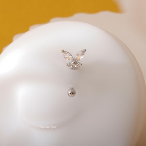 Puede incluir: Una joya de cuerpo de mariposa plateada con detalles de cristal transparente. La mariposa está unida a una barra plateada con una bola transparente en el extremo.