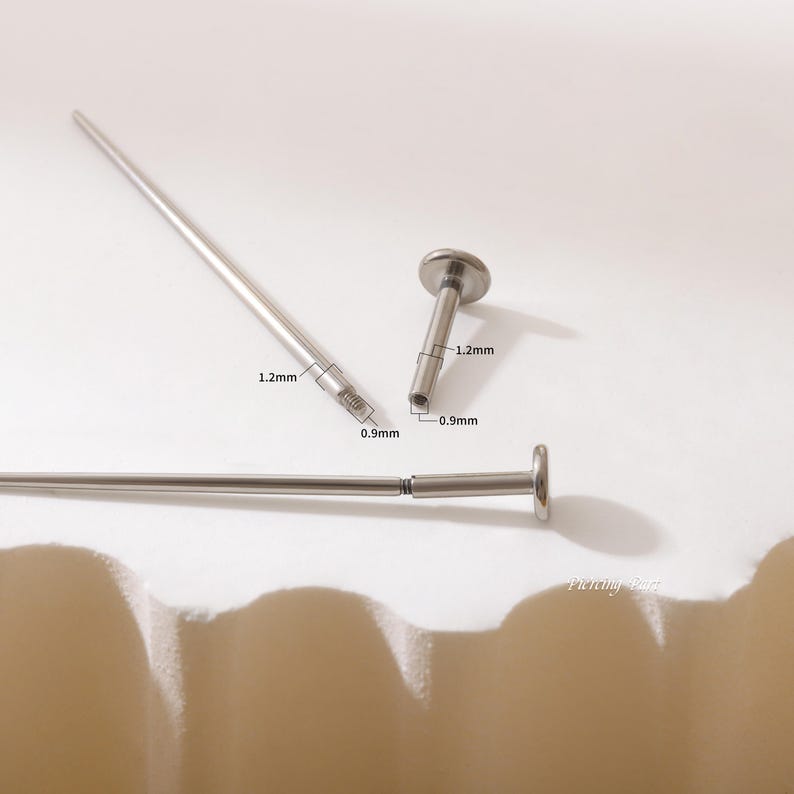 May include: Two silver barbell posts with a 0.9 mm diameter and 1.2 mm length. The posts are threaded and have a flat top.