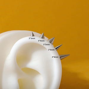 May include: A white ear model displays five silver-toned metal cone-shaped studs of varying sizes. The studs are labeled with dimensions: 3*3mm, 4*4mm, 5*5mm, 3*6mm, and 4*8mm, indicating the base diameter and height of each individual spike.