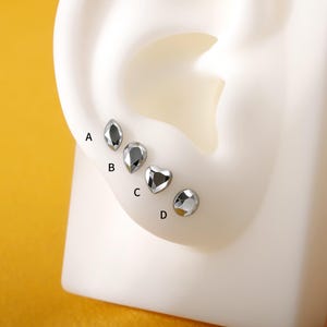 May include: Four silver-colored stud earrings in different shapes: a marquise, a teardrop, a heart, and a round shape. The earrings are displayed on a white ear model against a yellow background. Each earring is labeled with a letter.