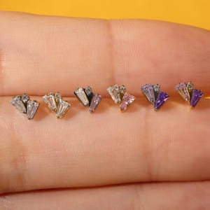 May include: A collection of six stud earrings, each featuring a fan-shaped design with baguette-cut and round-cut clear crystals. The earrings are in various colors, including clear, pink, and purple. The earrings are displayed on a yellow background.
