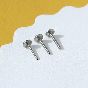 May include: Three silver body jewelry pieces, each with a flat circular top and a straight post. The pieces are arranged in order of size, with the smallest on the left and the largest on the right.