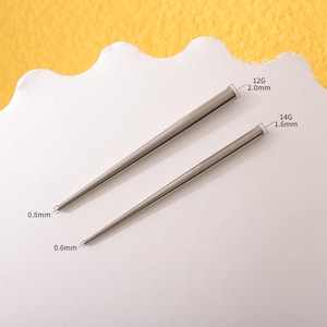 Puede incluir: Dos barras de piercing de metal plateado, una de calibre 14 y 1,6 mm de grosor, y la otra de calibre 12 y 2,0 mm de grosor. Las barras son rectas y tienen un extremo puntiagudo.