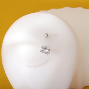 May include: A silver body piercing with a small, iridescent planet charm and two stars. The charm is attached to a silver ring.