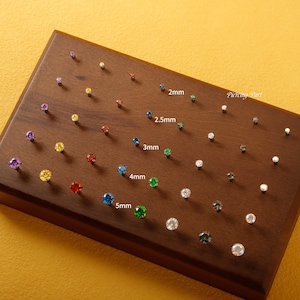 Peut inclure: Une collection de boucles d'oreilles à clous en pierres précieuses de différentes couleurs et tailles, présentées sur une surface en bois foncé. Les tailles sont indiquées de 2 mm à 5 mm. Les boucles d'oreilles comprennent des pierres précieuses violettes, jaunes, rouges, bleues et vertes.