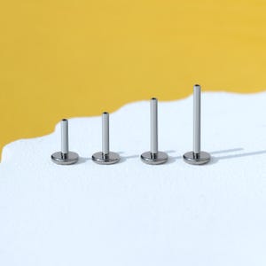 May include: Four silver barbell studs of different lengths, arranged in order of size from smallest to largest on a white surface.
