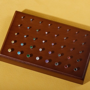 May include: A wooden display case filled with various small, colorful gemstone studs. The studs are set in silver-colored metal and arranged on a dark brown, rectangular surface. The gemstones are in a variety of colors, including yellow, blue, green, and purple.