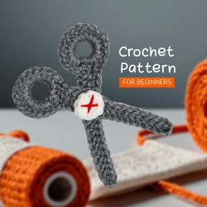 Pode incluir: Padrão de crochê para iniciantes. Um par de tesouras de crochê cinza com um botão branco e uma cruz vermelha.