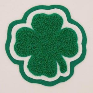 May include: A green, textured four-leaf clover patch with a white and green border. The clover is a symbol of good luck and is often associated with St. Patrick's Day. The patch is likely made of felt or a similar material.