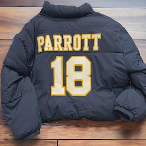 May include: A navy blue puffer jacket with the name "PARROTT" and the number "18" in yellow lettering. The jacket has a quilted design and a high collar. The jacket is lying on a wooden surface.