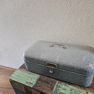 May include: A gray, rectangular metal box with a textured surface, a handle, and a latch. The box is sitting on a wooden surface with a distressed green painted section. The background is a textured, light gray wall.