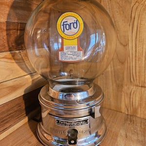 以下が含まれることがあります： 透明なガラスのグローブと銀色の金属ベースを備えた、ヴィンテージのフォードガムボールマシン。グローブにはフォードのロゴと製品情報があります。ベースにはコインスロットとディスペンシングメカニズムがあります。