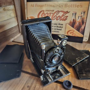 May include: A vintage black folding camera with a bellows design, lens, and various dials. The camera is on a wooden surface, with several black film holders nearby. A vintage Coca-Cola advertisement is in the background.