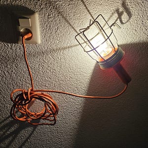 Puede incluir: Una lámpara de trabajo naranja con una jaula protectora de alambre está enchufada a una toma de corriente. La luz proyecta un brillo brillante y una sombra en la pared texturizada. El cable naranja está enrollado.