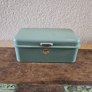May include: A vintage, rectangular bread box in a muted teal color. It features a curved lid, a silver handle, and a small, circular emblem on the front. The bread box is made of metal and sits on a wooden surface.