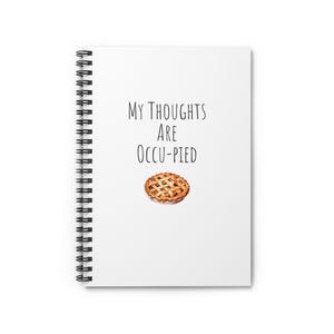 May include: A white spiral-bound notebook featuring the black text "MY THOUGHTS ARE OCCU-PIED" centred on the front cover, positioned above a small, detailed illustration of a golden-brown lattice-crust fruit pie.