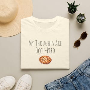 May include: A cream-coloured t-shirt featuring the black text "MY THOUGHTS ARE OCCU-PIED" printed above a small, detailed illustration of a golden-brown lattice-crust pie. The shirt has a simple crew neckline and a clean, minimalist design.
