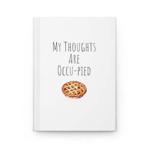 May include: A white hardback notebook featuring the black text 'MY THOUGHTS ARE OCCU-PIED' centred on the front cover, positioned above a small, detailed illustration of a golden-brown lattice-crust fruit pie.