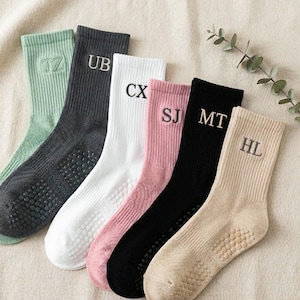 Könnte beinhalten: Sechs Paar Crew-Socken in verschiedenen Farben: Hellgrün, Dunkelgrau, Weiß, Rosa, Schwarz und Beige. Jede Socke hat Initialen auf dem Bündchen gestickt. Die Sohlen haben einen rutschfesten Grip.