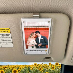 Peut inclure: Un cadre photo en acrylique transparent fixé à un pare-soleil de voiture, affichant une photo de mariage d'un couple. La photo est prise devant un mur de briques rouges. Le texte "Our Adventure Begins" est imprimé en bas.