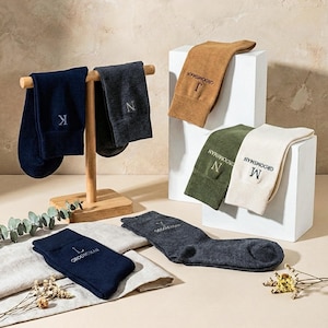 Puede incluir: Una variedad de calcetines personalizados en varios colores, incluyendo azul marino, gris, marrón, verde oliva y crema. Los calcetines están bordados con iniciales y la palabra "Groomsman". Algunos calcetines se exhiben en un soporte de madera y bloques blancos.