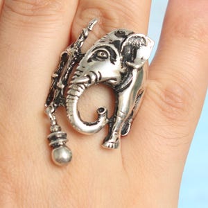 May include: A silver-colored ring featuring an elephant design. The ring has intricate details, including the elephant's trunk, tusks, and ears. A small, decorative element hangs from the side of the elephant. The ring is worn on a finger.