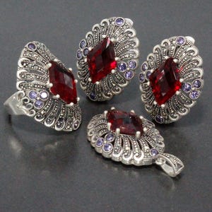 May include: A jewelry set featuring a ring, earrings, and a pendant. Each piece has a large, faceted, red gemstone at the center, surrounded by smaller purple stones and a silver-colored, ornate setting. The set is displayed on a dark surface.