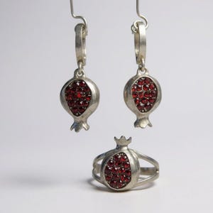 Può includere: Set di gioielli color argento con design a melograno. Il set include un anello e un paio di orecchini. Ogni pezzo ha la forma di un melograno con un gruppo di pietre preziose rosse. Gli orecchini hanno una chiusura a gancio.