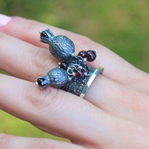 May include: A silver ring featuring a pomegranate design. The ring has two textured pomegranate fruit with small red beads. The ring is worn on a finger, with a blurred green background.