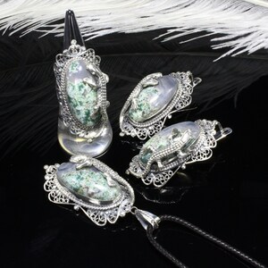 May include: A silver jewellery set comprising a ring, two earrings, and a pendant. Each piece features an oval stone with green and grey patterns, and a silver alligator design. The ring is tall, and the pendant hangs from a black cord.