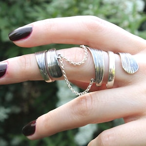 Könnte beinhalten: Ein silbernes Mehrfinger-Ring-Set mit einer Kette. Das Ring-Set umfasst mehrere Bänder, die durch eine zarte Kette verbunden sind. Die Ringe haben ein strukturiertes, blattartiges Design. Die Nägel sind dunkelburgunderfarben lackiert.