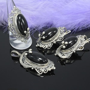 May include: A jewellery set comprising a ring, pendant, and two earrings. Each item features a large, oval, glossy black stone set in a silver filigree frame. The ring is displayed on a clear stand. The earrings have a hook closure.