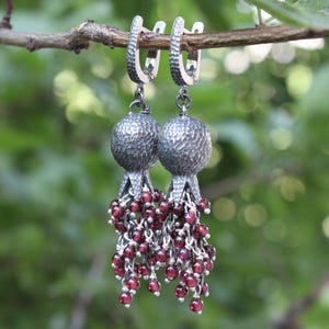 May include: A pair of silver earrings with a textured, spherical design, each adorned with a cascade of small, red beads. The earrings are suspended from silver hoops and are displayed against a blurred green background.