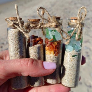 May include: Four small glass bottles with cork tops, tied with twine, filled with sand and sea glass. The bottles contain different colours of sea glass, including green, amber, and clear.