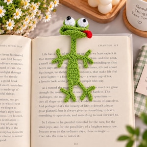 Peut inclure: Un marque-page en crochet vert en forme de grenouille avec de grands yeux blancs, des pupilles noires et une petite langue rouge tirée. La grenouille a un long corps avec quatre membres écartés et est posée sur les pages d'un livre ouvert.