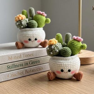 May include: Two crocheted cactus plants in white pots with smiling faces and small brown feet. Each pot contains green, yellow, and pink yarn cacti with white bead accents. The pots are placed on a wooden surface next to a stack of three books.