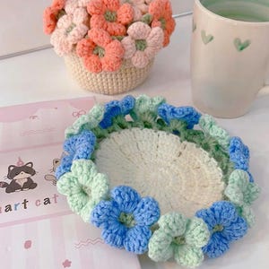 以下が含まれることがあります： 青と緑の花柄デザインの手作りかぎ針編みコースター。ピンクとオレンジの花が入ったお揃いのかぎ針編みフラワーバスケットが近くにあります。緑のハートのディテールが入った白いマグカップも写っています。