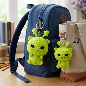 Könnte beinhalten: Zwei hellgrüne gehäkelte Alien-Schlüsselanhänger mit schwarzen Augen und Antennen sind an einem blauen Rucksack und einer beigefarbenen Tragetasche befestigt. Die Schlüsselanhänger sind handgefertigt und haben ein skurriles Design.