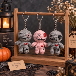 May include: Three crochet voodoo doll keychains hanging from a wooden stand. Two dolls are grey and one is light pink. Each features a stitched mouth, one button eye, one X-stitched eye, and a small red heart patch. A sign reads Small Crochet Big Happiness.